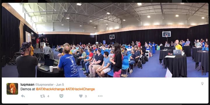 wide angle photo of a gym full of people at a hackathon event listenting to presentations from a stage